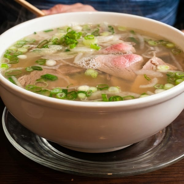 Steaming bowl of pho with beef, noodles, and green onions on a wooden table.
