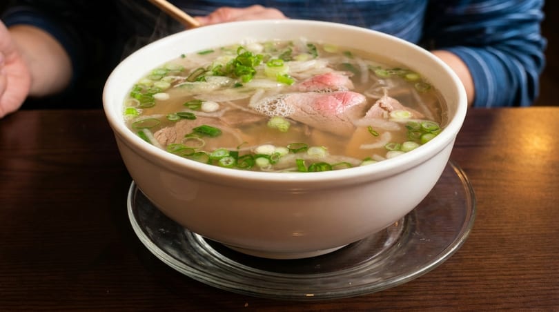 Steaming bowl of pho with beef, noodles, and green onions on a wooden table.
